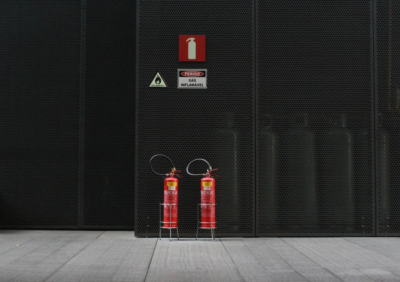 Dois extintores junto a uma parede escura, sob placas de sinalização de extintor e de gás inflamável